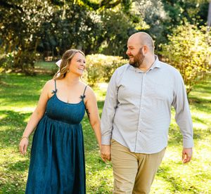 A couple walks together through a sun-dappled garden path in casual attire.