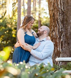 A romantic couple embraces by a large tree trunk in a sunlit forest setting.