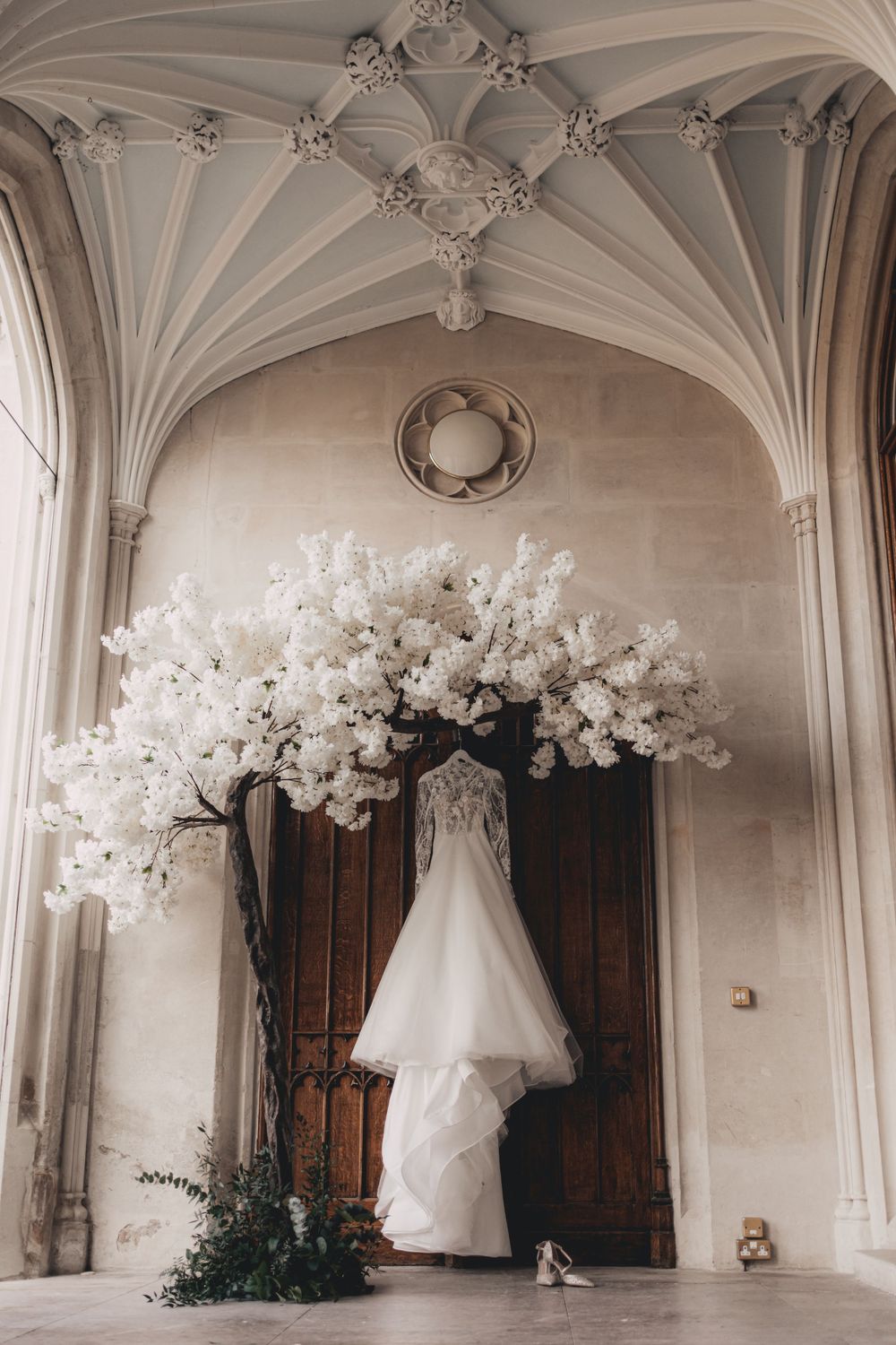 Wedding dress hanging under ornate vaulted ceiling with white floral arch display at Ashridge House.
