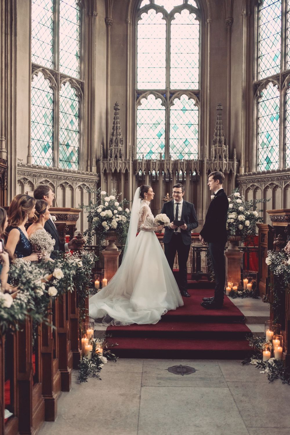 A wedding ceremony in a gothic cathedral with ornate windows and floral decorations lining the aisle at Ashridge House.