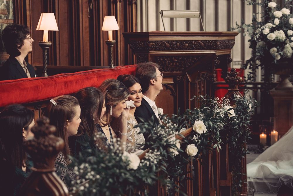 Wedding guests seated in wooden pews decorated with floral arrangements and candles at Ashridge House.