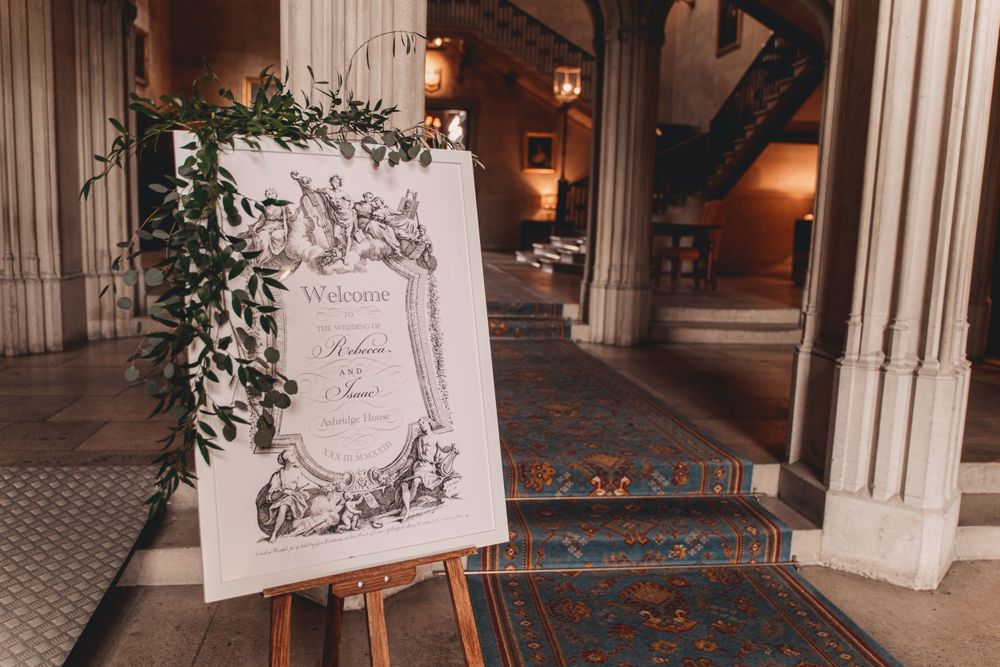 Ornate welcome sign with floral illustration displayed on easel at wedding entrance at Ashridge House.