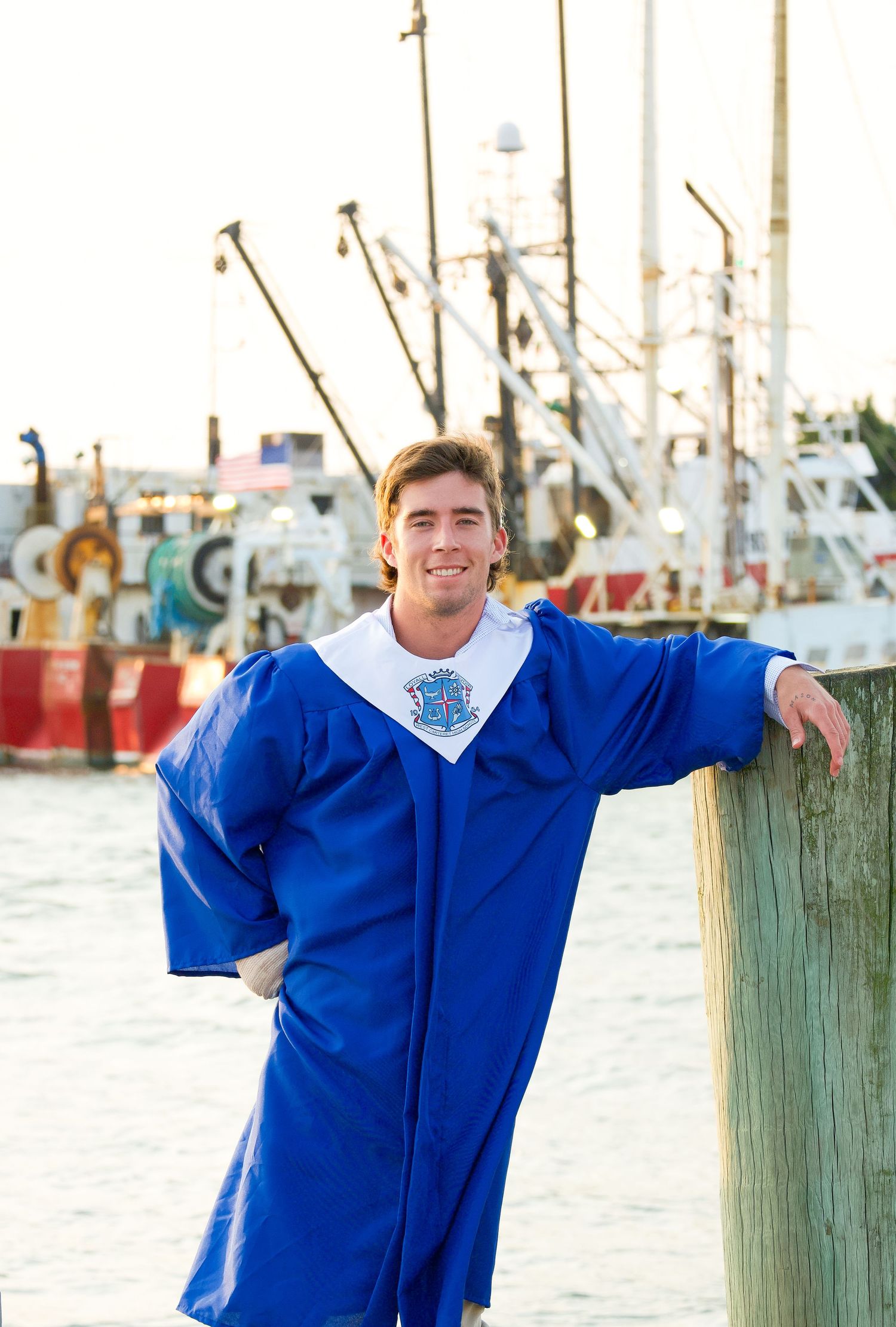 A person in a blue graduation gown poses by a dock with fishing boats in background.