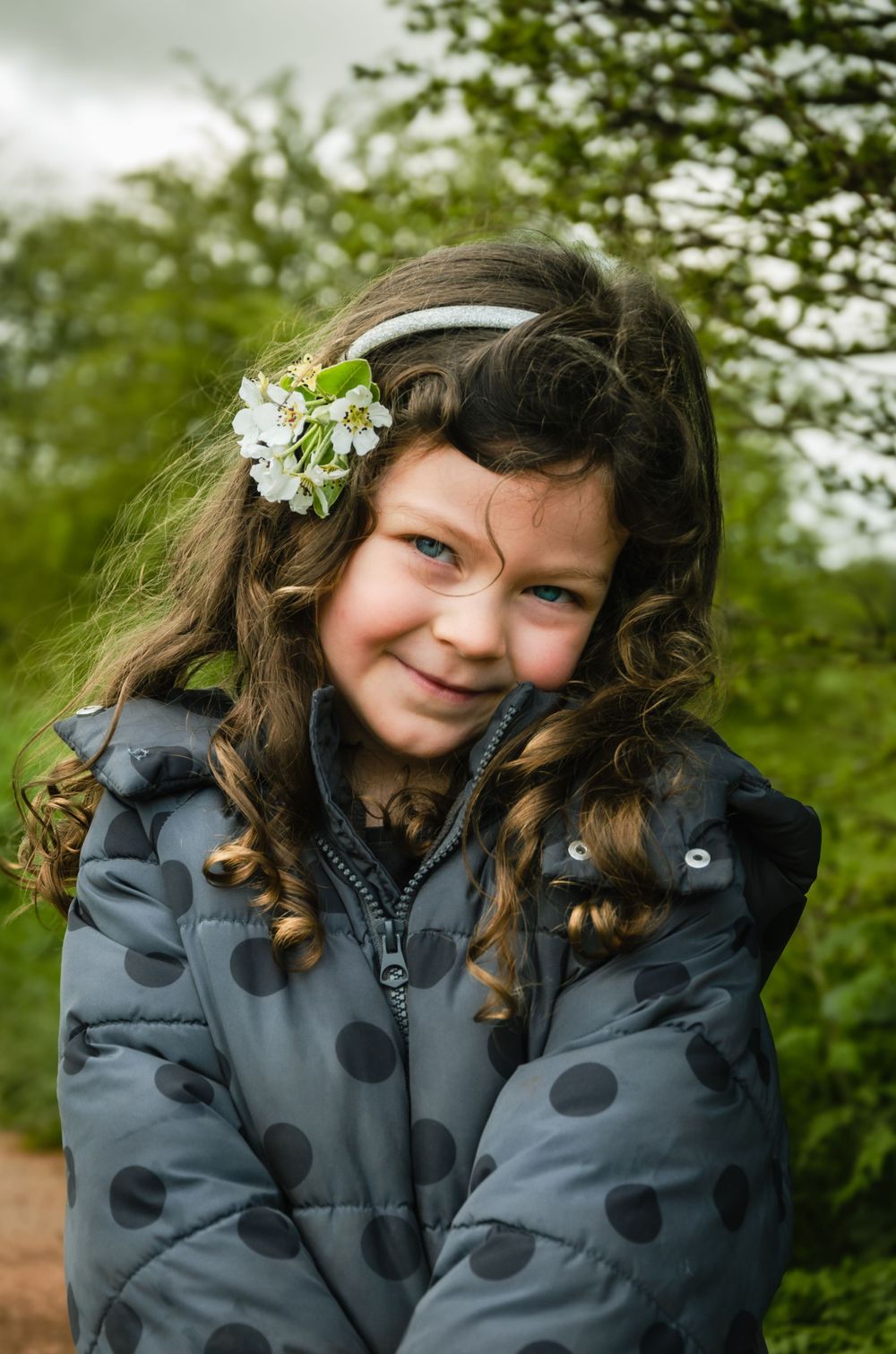 Child in polka dot jacket with flower accessory smiling in natural outdoor setting.