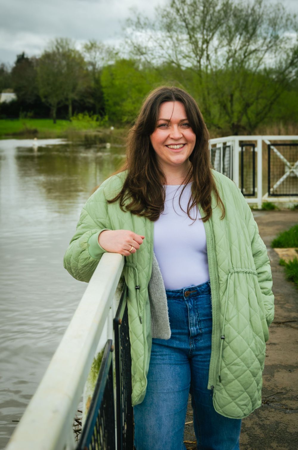 Woman in green quilted jacket smiling while standing on dock near water.