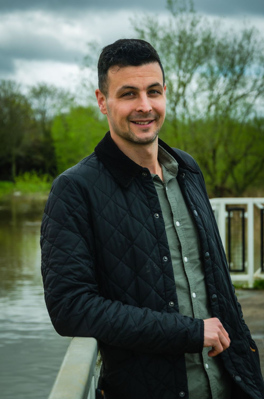 Man in black quilted jacket smiling on dock with water and trees in background.