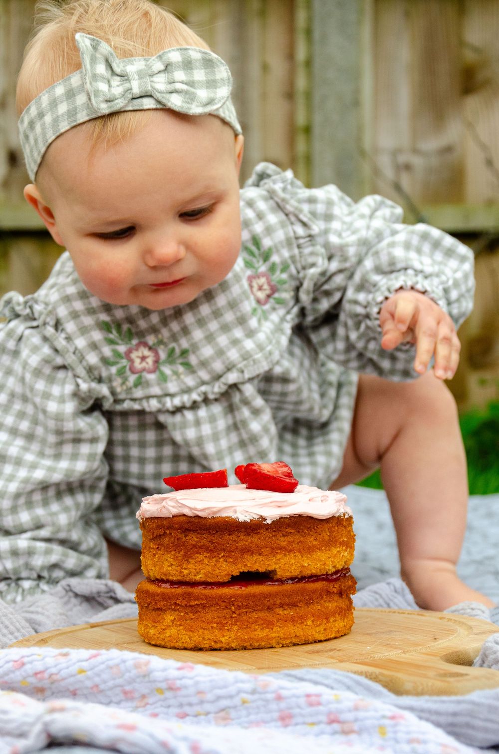 A small child in a gingham dress and matching headband looks down at a layered cake with strawberries on top.