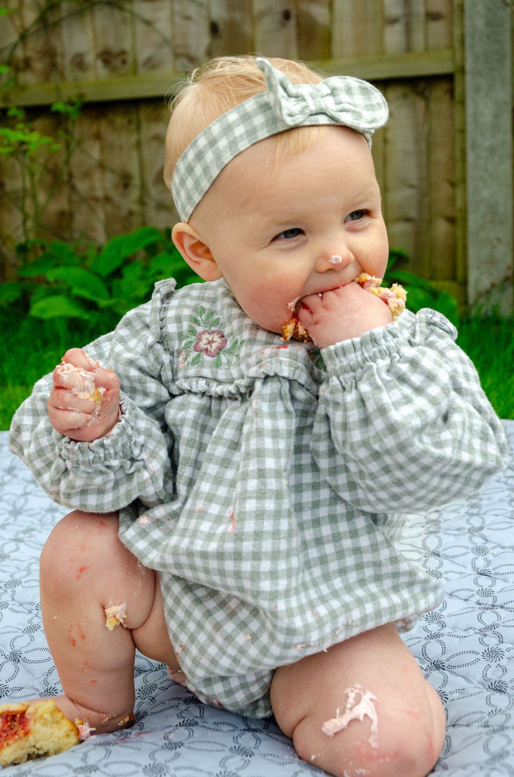 A baby in a gray gingham outfit and matching headband sits outdoors on a blanket while eating.