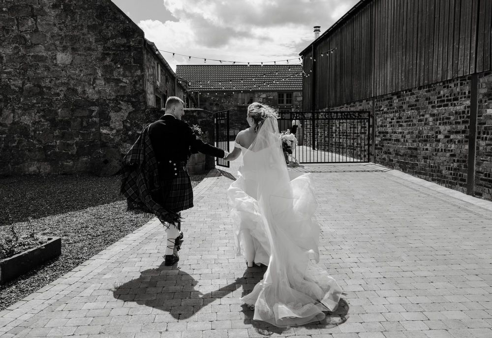 A couple dances joyfully together in a Scottish courtyard, with the bride's dress and veil flowing.