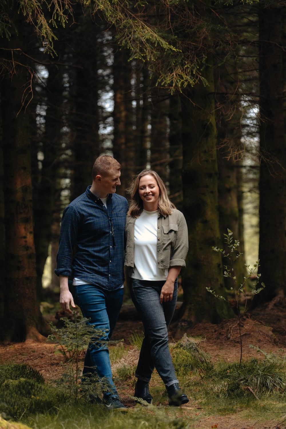 A couple walks and laughs together through a lush forest, wearing casual jeans and jackets.
