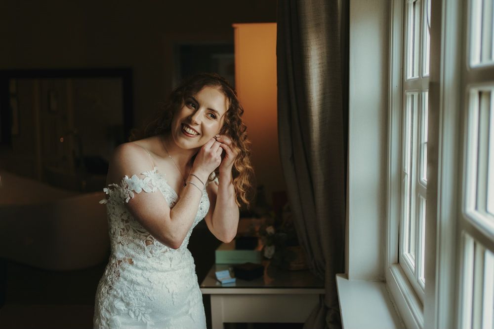 Bride in lace off-shoulder wedding gown smiling while putting on earrings near a bright window.