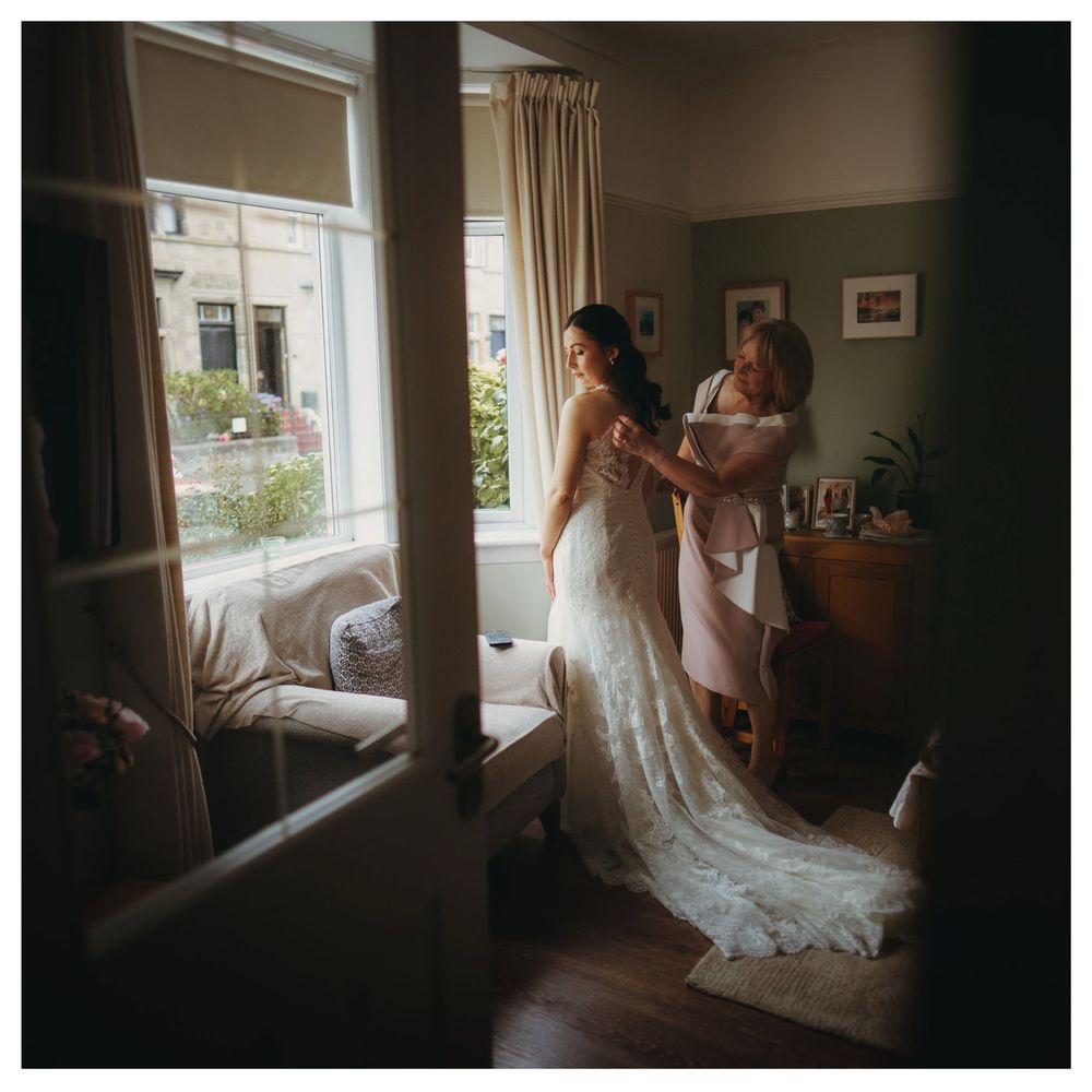 A bride in a white lace gown being helped by a woman near a sunlit window in an elegant room.