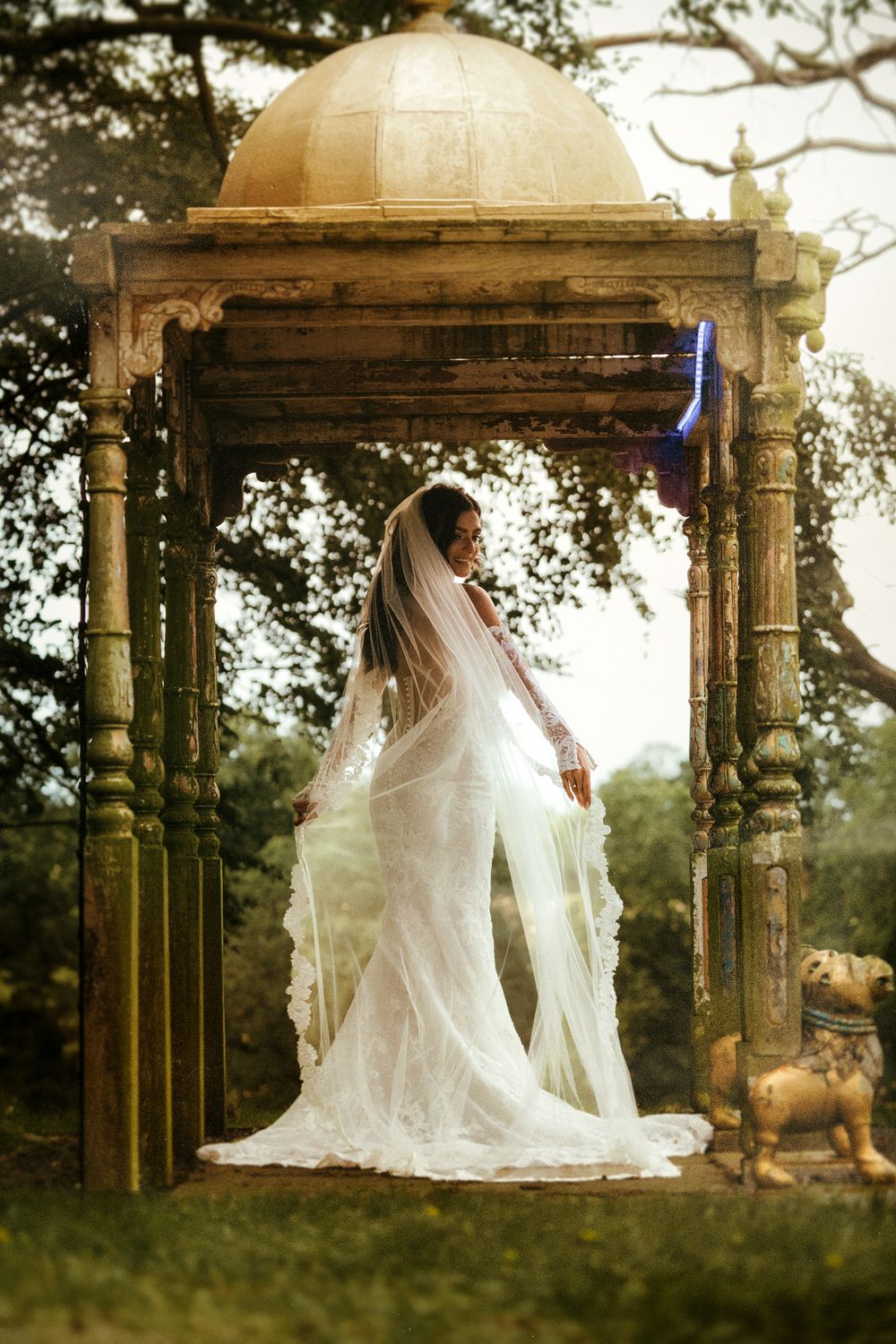 Bride in white lace gown and veil stands under ornate stone gazebo surrounded by lush green trees.
