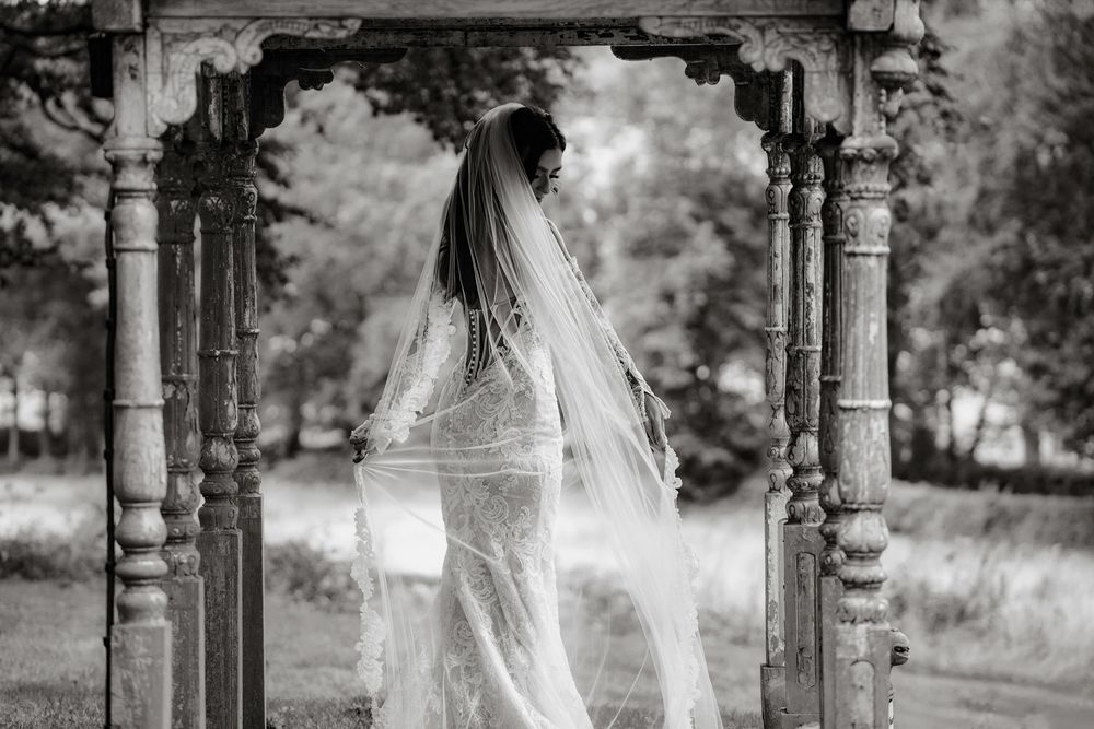 Black and white photo of a bride in a lace gown and veil standing under a stone archway outdoors.