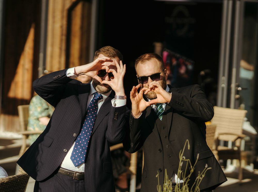 Two people in suits making silly gestures while taking photos outdoors.