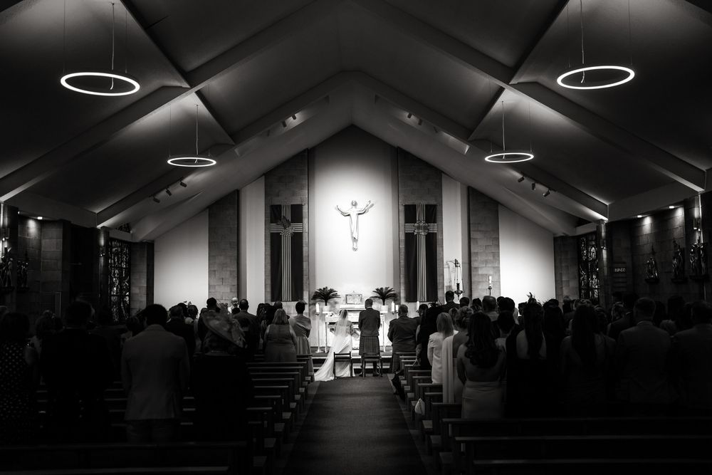 Black and white photo of a wedding ceremony inside a church with guests seated in pews and a cross above the altar.
