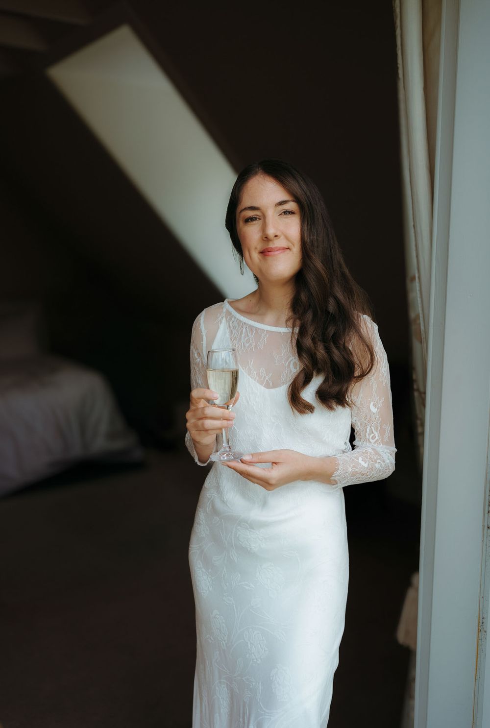 Bride in white lace gown holding champagne glass, smiling indoors during wedding preparations.