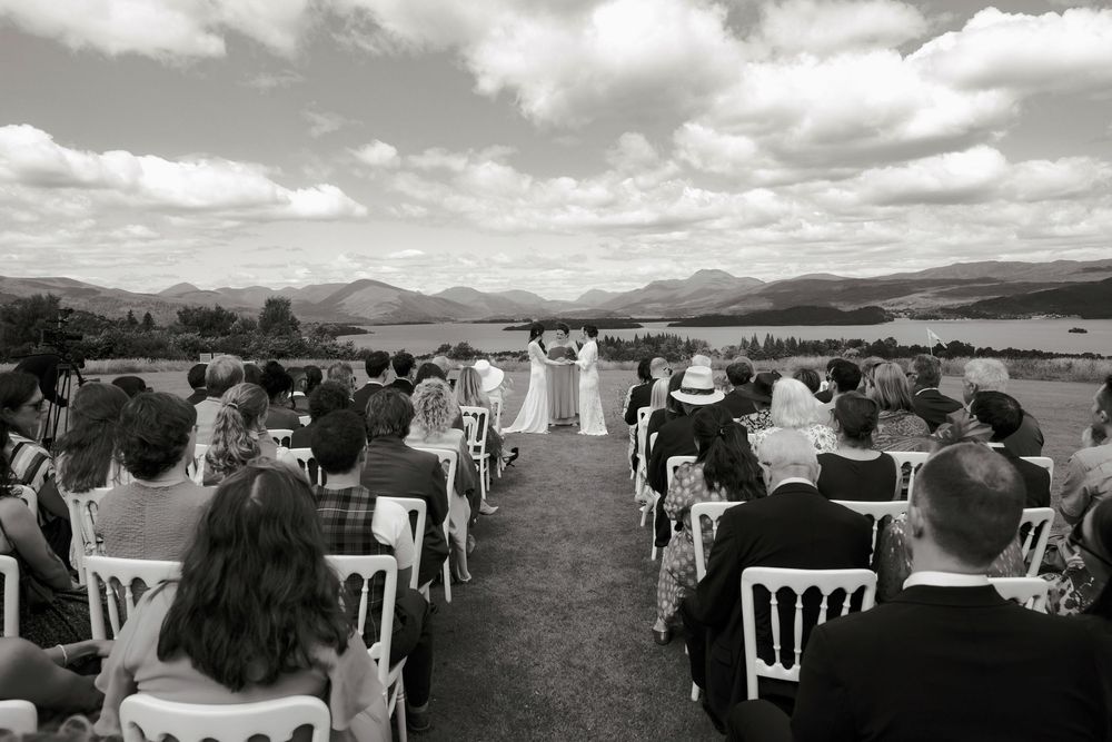 Black and white outdoor wedding ceremony with guests seated in white chairs, mountains and lake in the background.