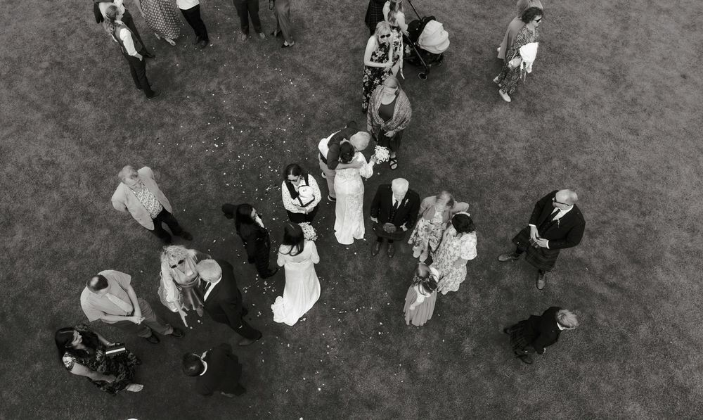 Aerial black and white photo of wedding guests gathered on grass around bride and groom.