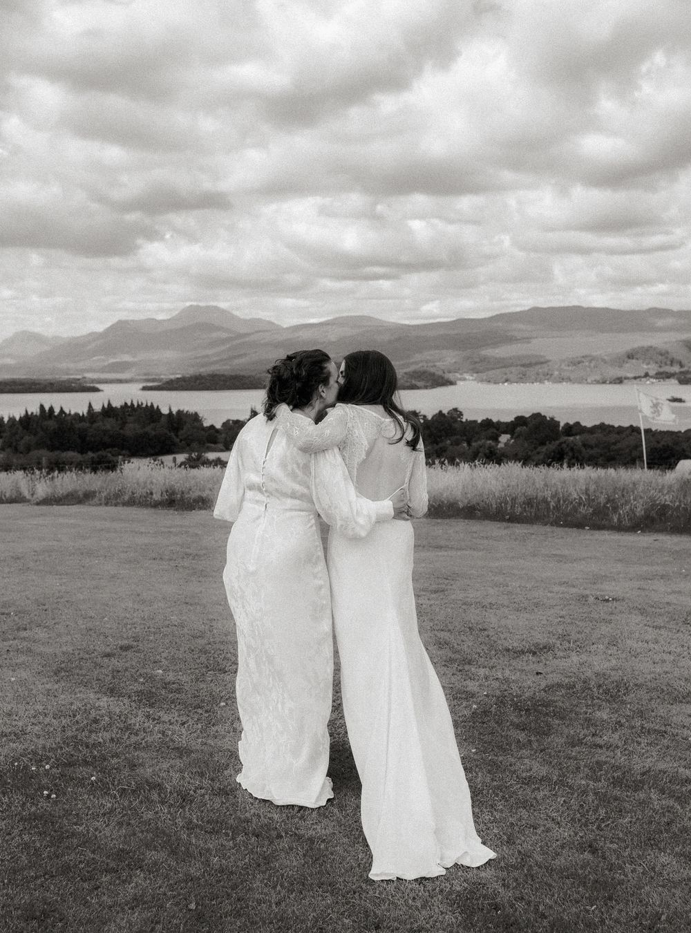 Two brides in white gowns embrace outdoors with scenic loch and mountain landscape in black and white wedding photo.