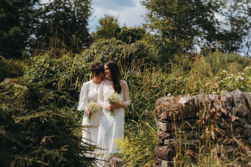 Two brides in white gowns holding bouquets stand together surrounded by lush greenery on their wedding day.