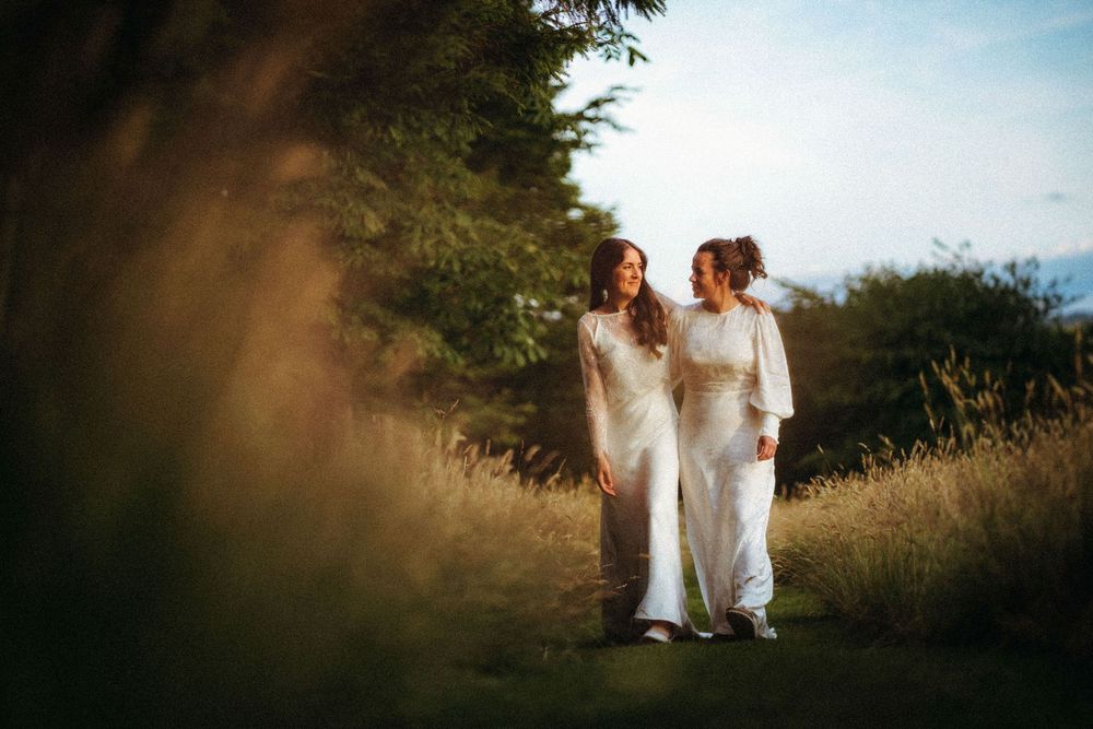 Two brides in white gowns walk together through a golden sunlit meadow, sharing a romantic glance.