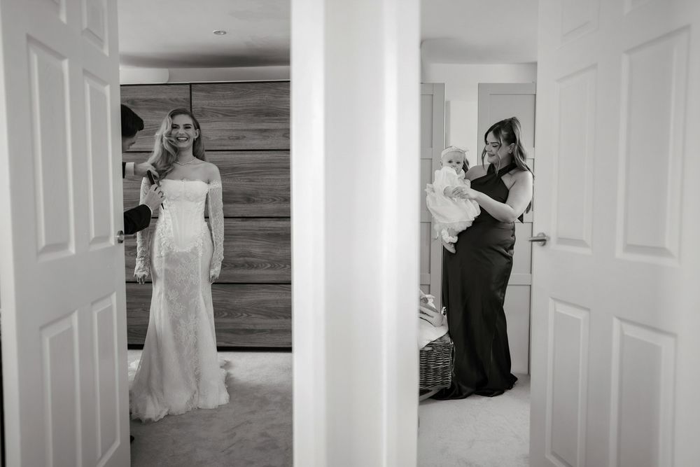 Black and white photo of a bride in a wedding gown and a woman holding a baby, captured through open doorways.