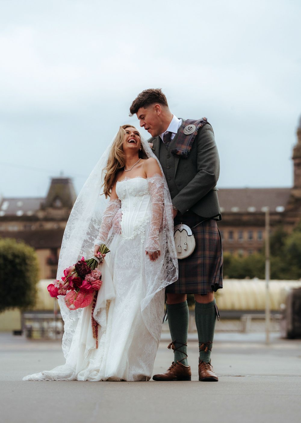 Bride in lace wedding dress holding pink bouquet laughs with groom in Scottish tartan kilt outdoors.