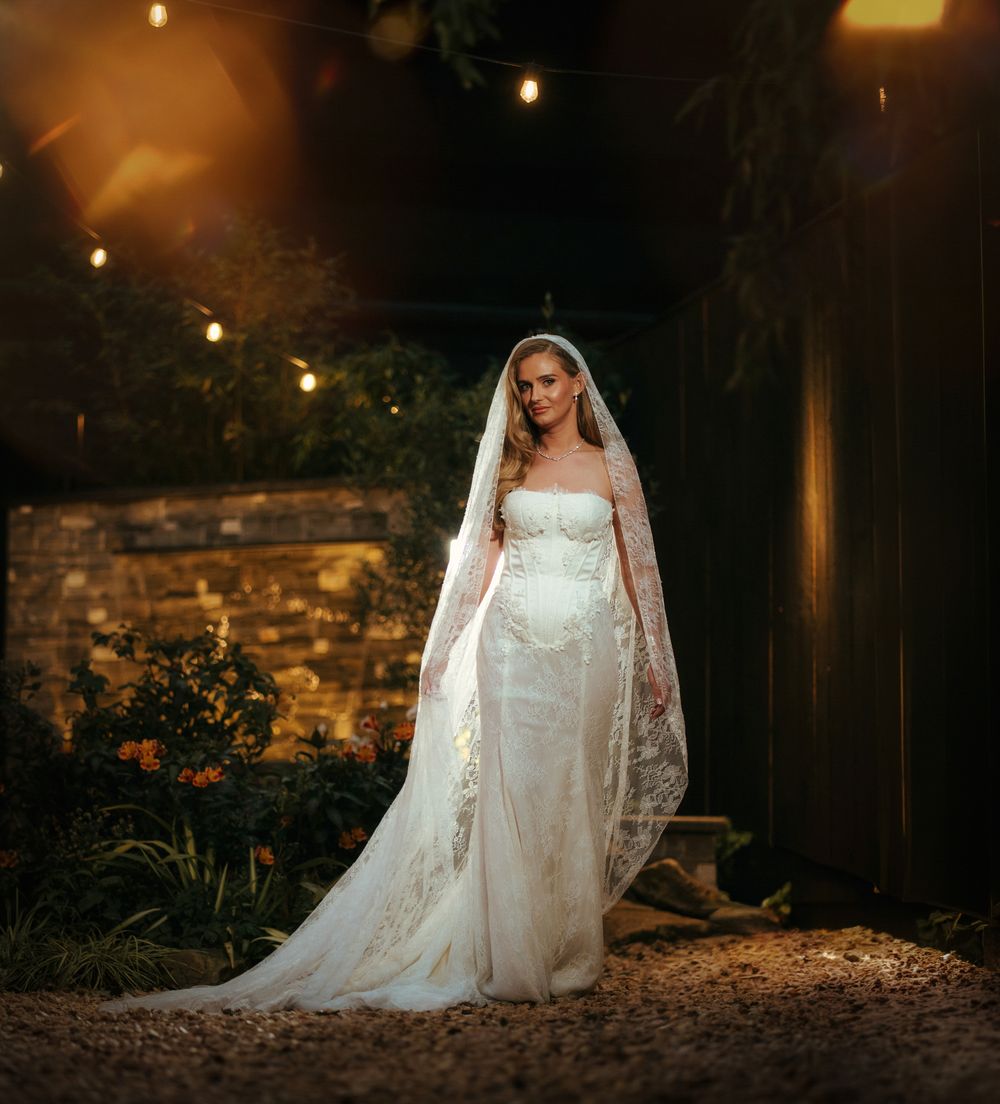 Bride in strapless white gown with long lace veil stands in warmly lit outdoor garden setting at night.