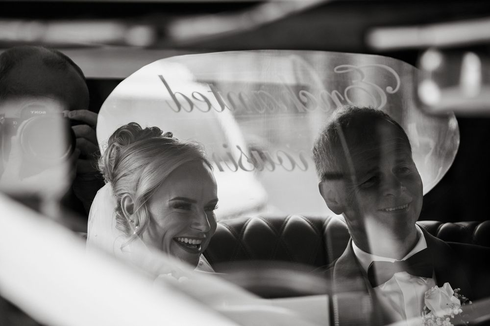 Black and white photo of smiling bride and groom seen through car window with decorative text reflection.