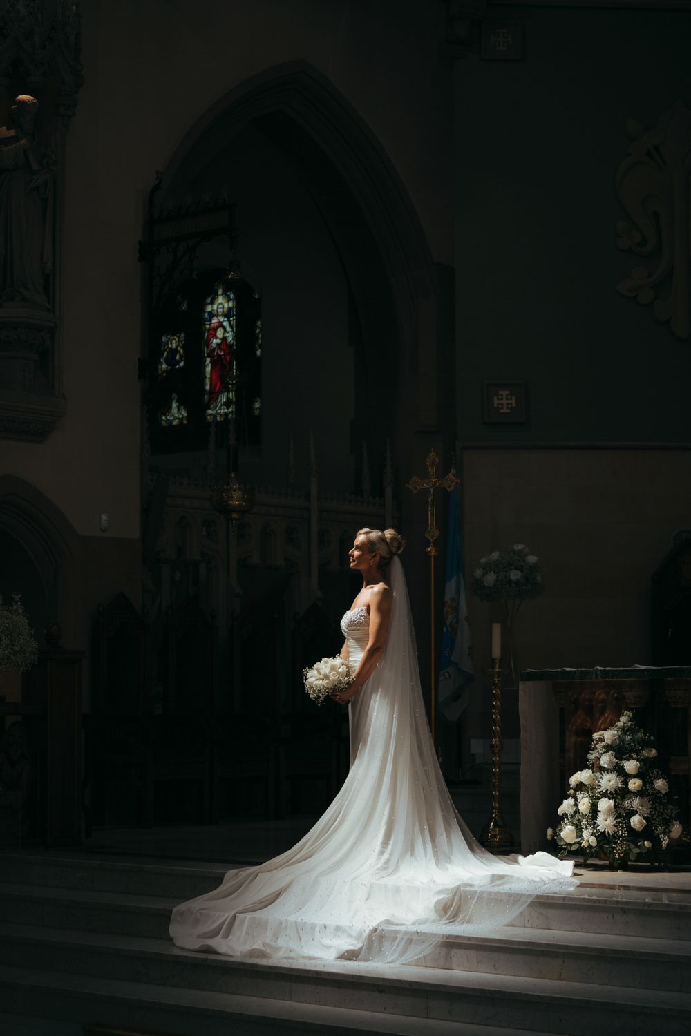Bride in white gown with veil holding bouquet stands at church altar with stained glass windows and floral arrangements.