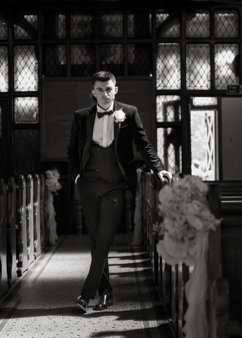 Groom in black tuxedo walks down church aisle with floral decorations in black and white wedding photo.