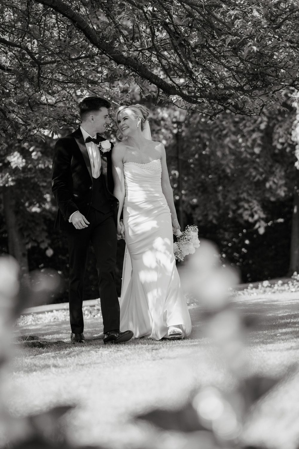 Black and white photo of a bride and groom walking together under blooming trees in a sunlit garden.