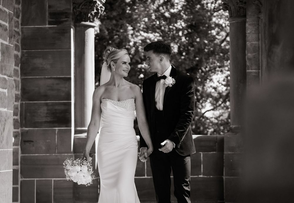 Black and white wedding photo of bride in strapless gown and groom in tuxedo holding hands near stone columns.