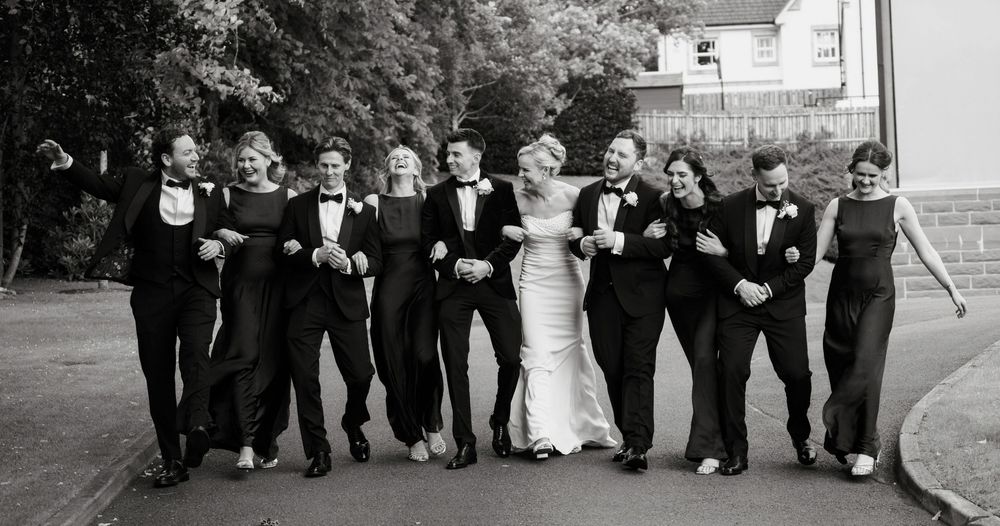 Wedding party in black tie attire walking together outdoors, bride in white gown surrounded by groomsmen and bridesmaids.