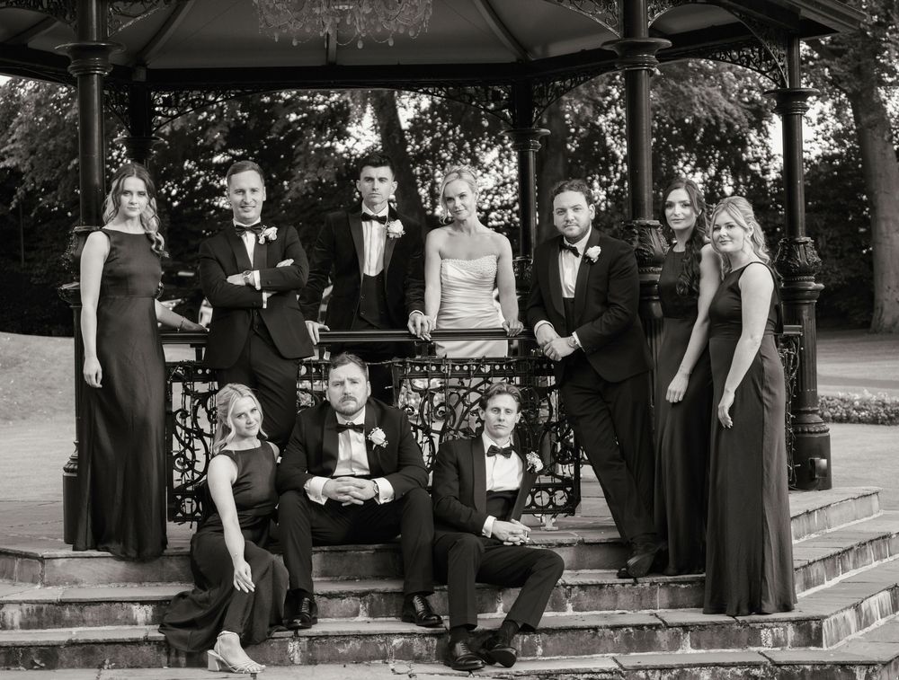 Black and white wedding party photo with bridesmaids and groomsmen posing on steps near a gazebo.