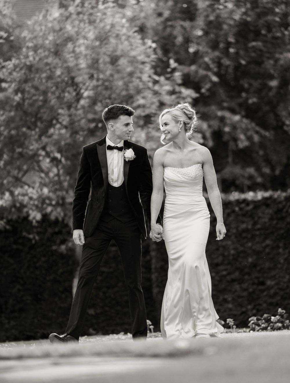 Black and white photo of a bride and groom walking hand in hand outdoors, smiling at each other on their wedding day.