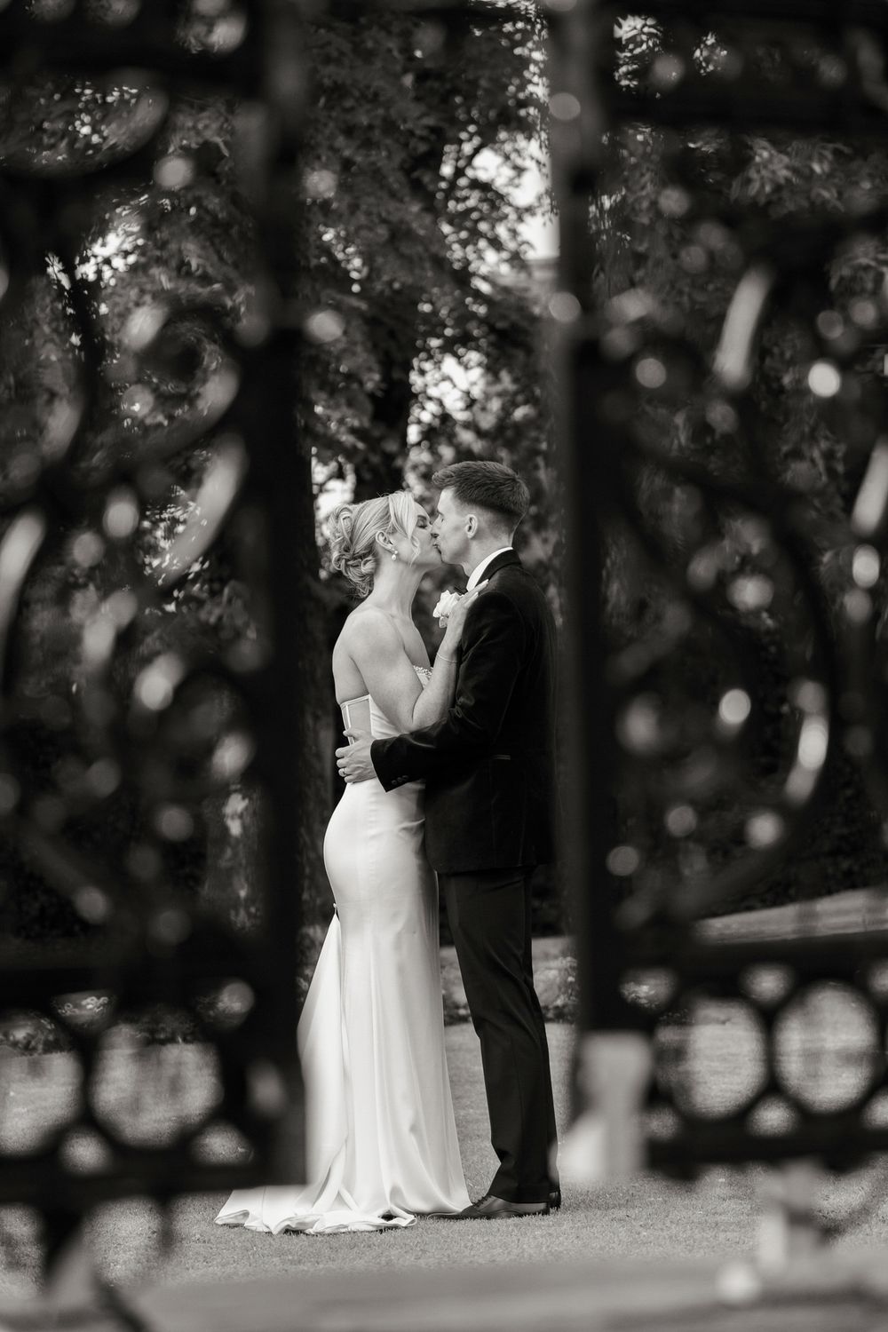 Black and white photo of a bride and groom sharing a kiss framed by ornate iron gates.