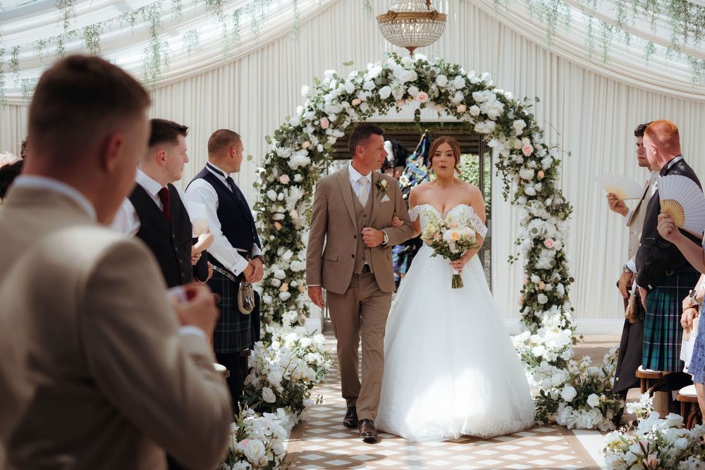 Bride and groom walk down the aisle under a white floral arch at an elegant wedding ceremony.