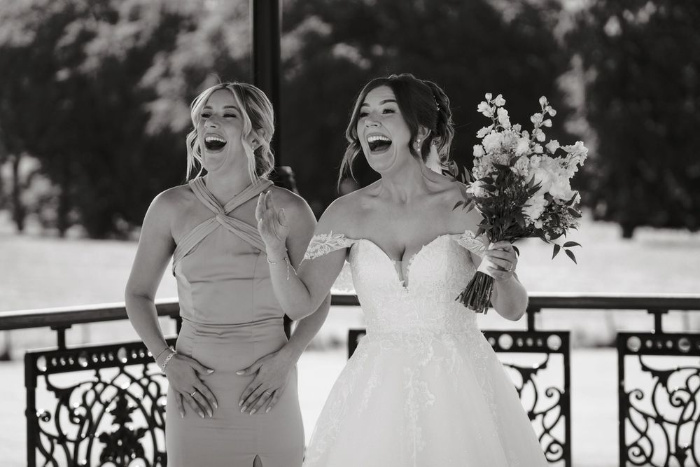 Bride and bridesmaid laughing joyfully outdoors, bride holding bouquet in elegant strapless gown, black and white wedding photo.