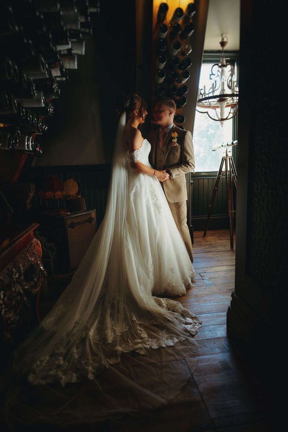 Bride in lace gown with veil and groom in tan suit share intimate moment in rustic dimly lit wedding venue.