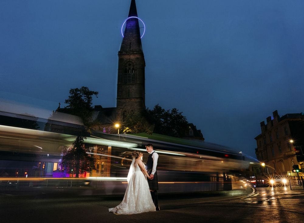 Bride and groom pose at night near illuminated church spire with light trails from passing tram in city street.