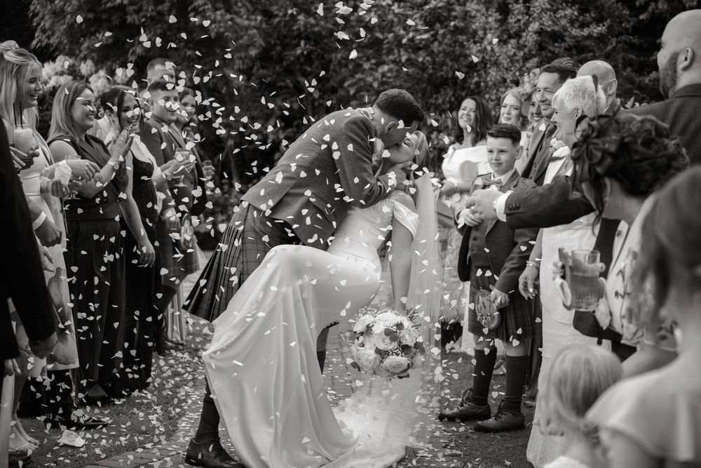 Black and white photo of newlyweds kissing amid confetti shower, surrounded by cheering wedding guests outdoors.