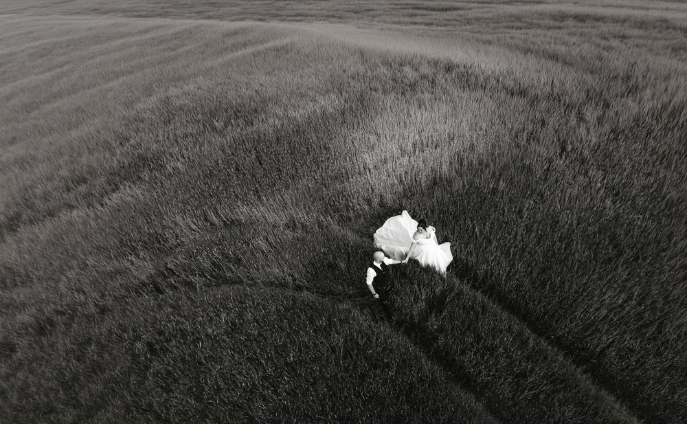 Aerial black and white photo of a person in white dress lying in vast grass field.