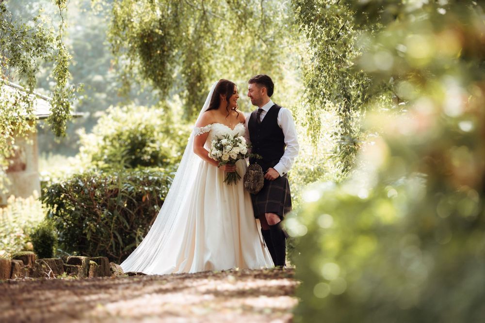 Bride and groom share a romantic moment in a lush green garden, with bride in white gown and groom in Scottish kilt.