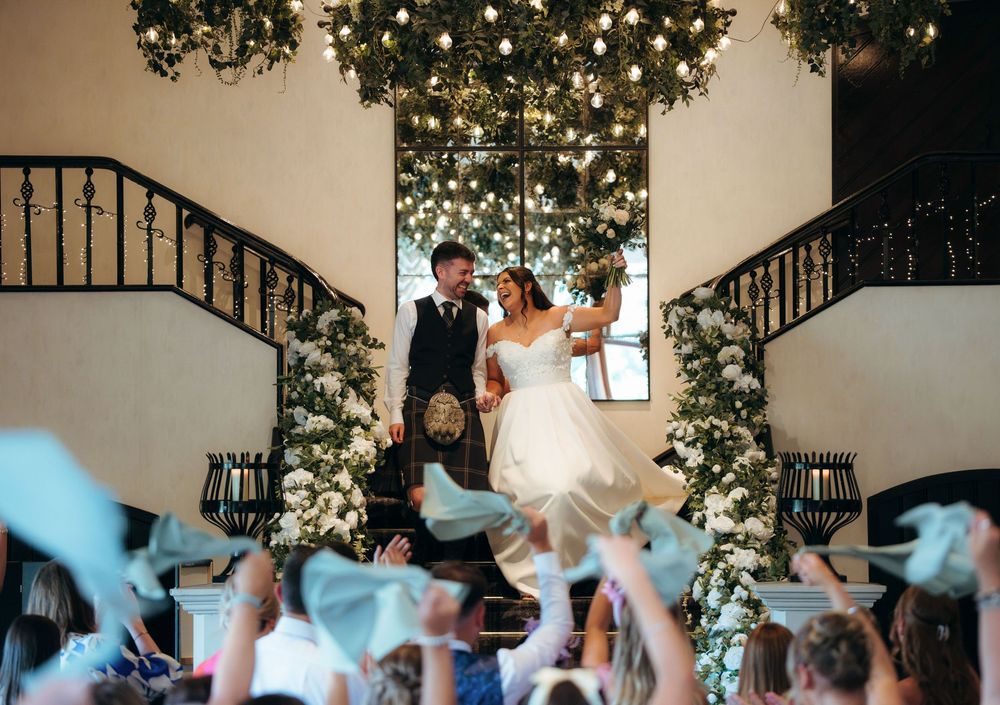 Bride and groom stand on grand staircase adorned with florals as guests wave ribbons at elegant wedding reception.