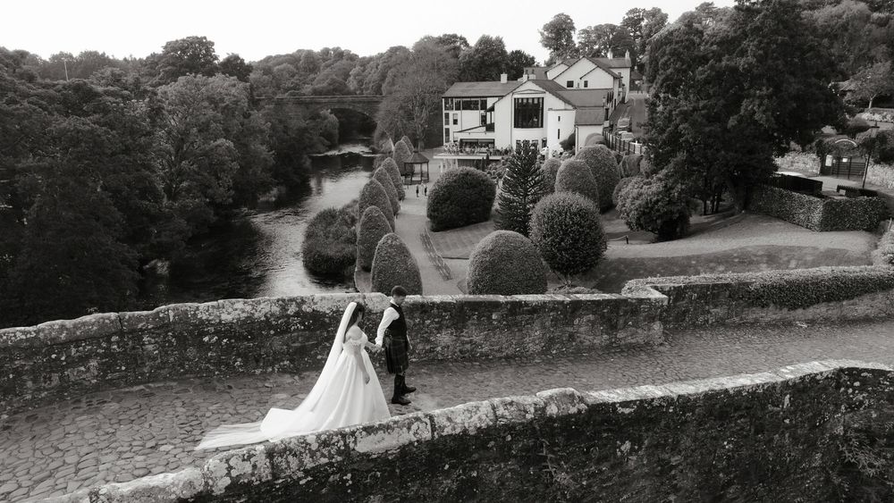 Black and white wedding photo of bride and groom on stone wall with manicured gardens and historic manor house backdrop.