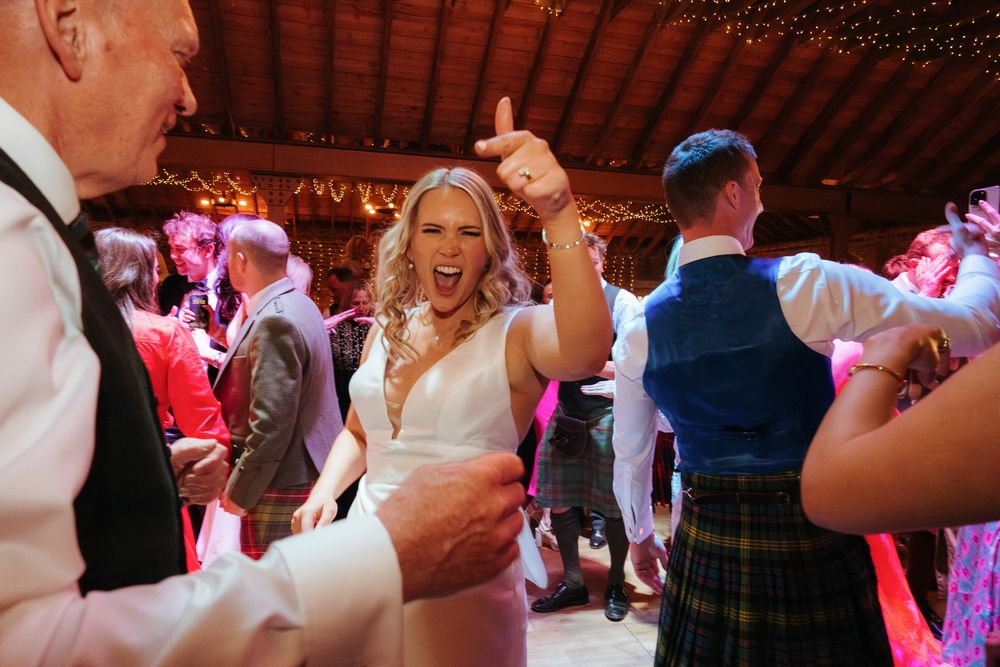 Bride dancing joyfully at wedding reception, pointing up, surrounded by guests in kilts and formal wear.