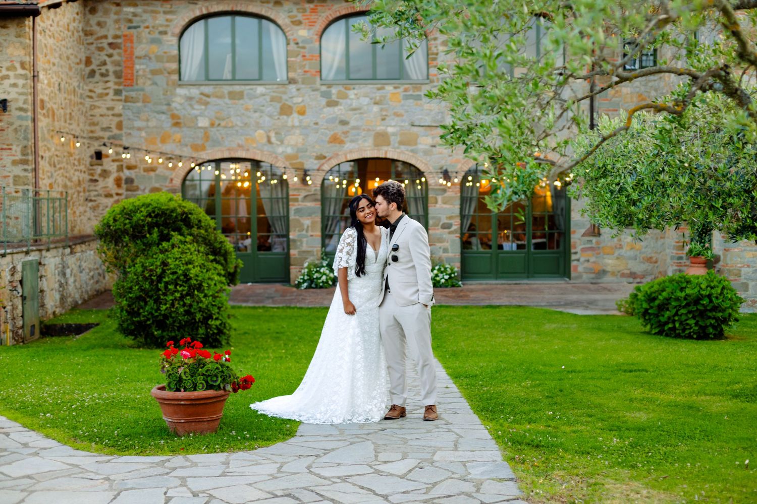 A couple poses in wedding attire outside a rustic stone building with arched windows.