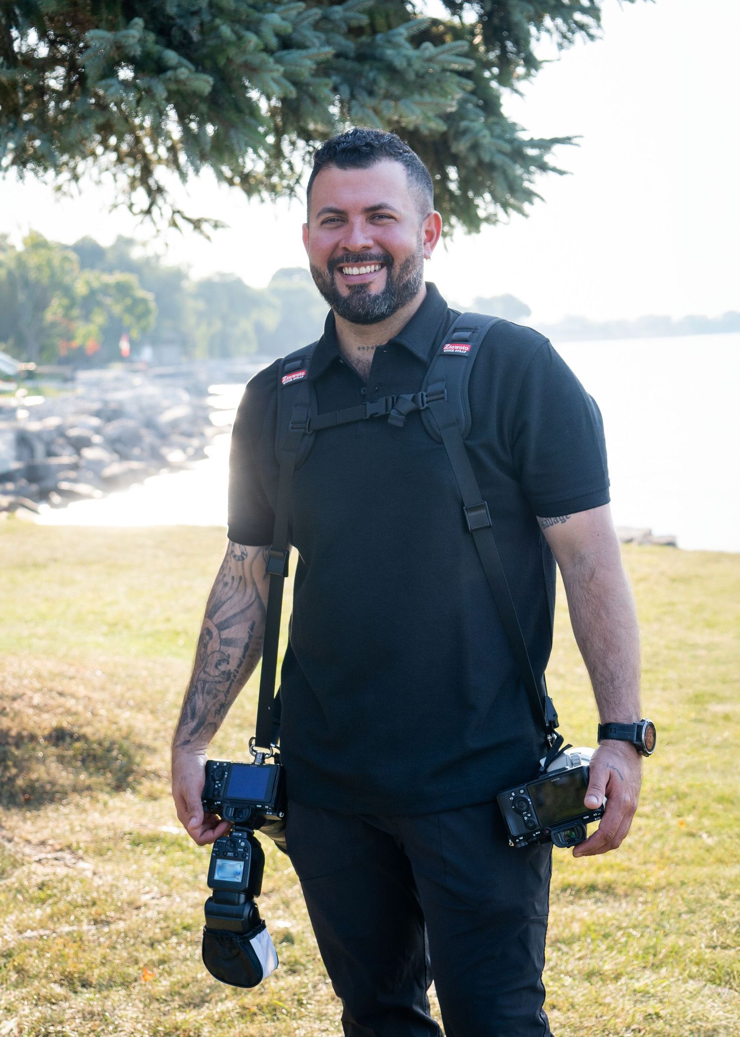 Professional photographer dressed in black holding two cameras stands on grass with evergreen trees and water in background.